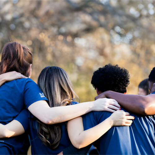 A group of individuals wearing matching shirts, photographed from the back with hands placed on each other's shoulders.