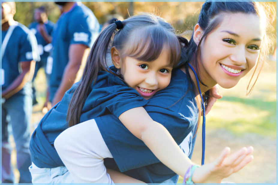A young girl, wearing the same uniform as a young adult, is carried on the adult's back while both look towards the camera with smiles, participating in a fundraising event.