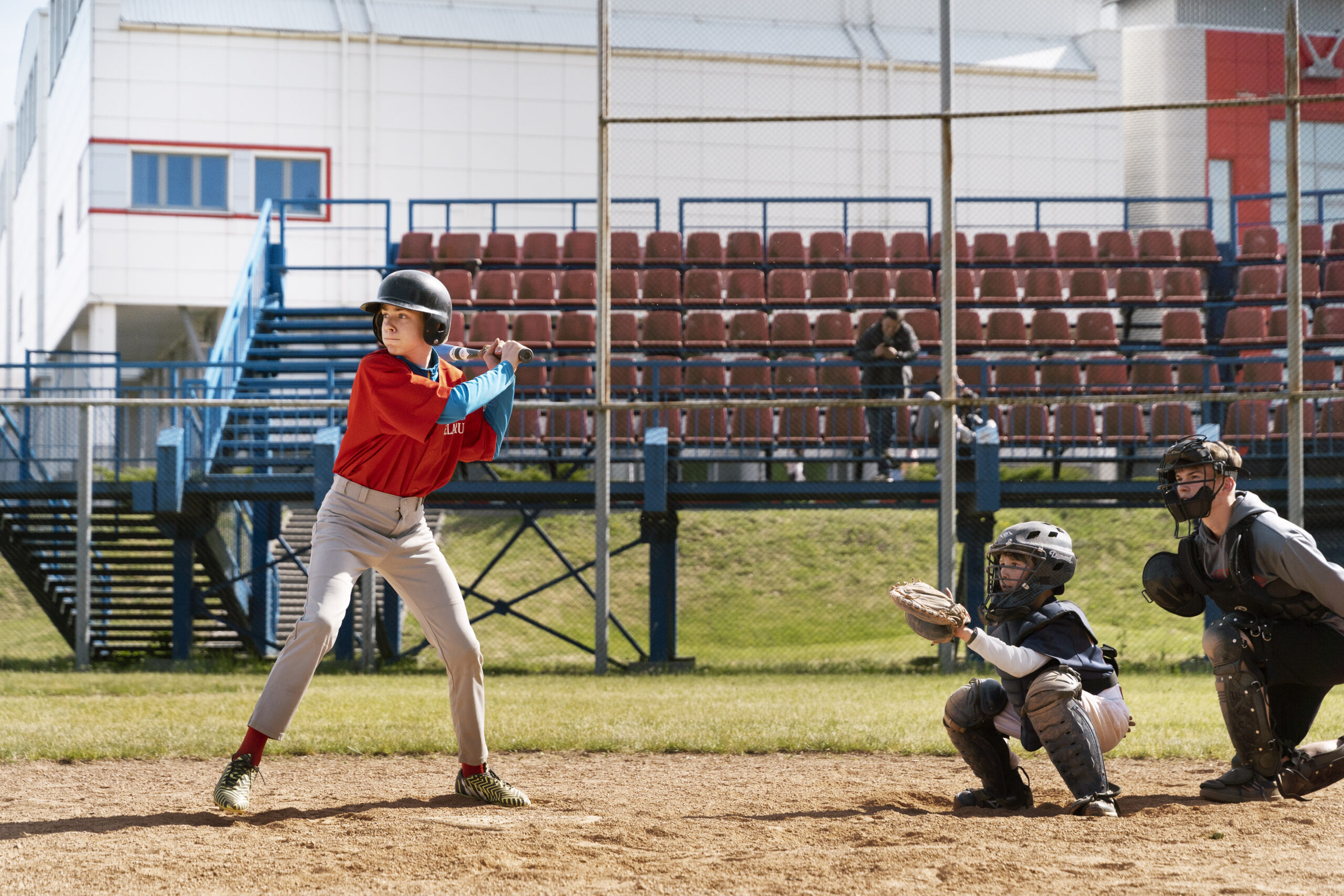 ChatGPT A young baseball player in a red jersey and helmet prepares to bat while the catcher and umpire crouch behind home plate on a sunny day.