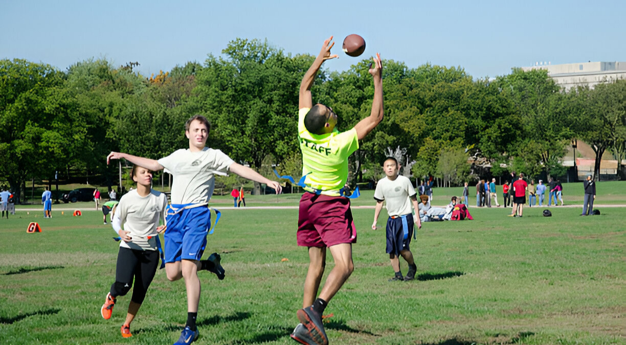 A group of young people playing flag football on a green field, with one player in a yellow shirt jumping to catch the ball.