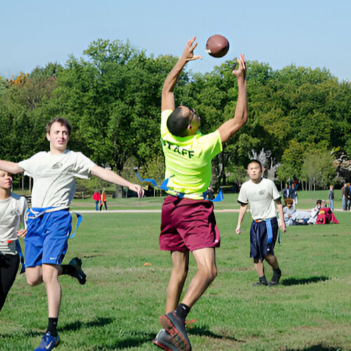 A group of young people playing flag football on a green field, with one player in a yellow shirt jumping to catch the ball.